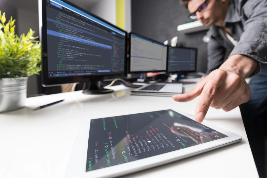 Man at computer desk with code on screens and reaching for a tablet